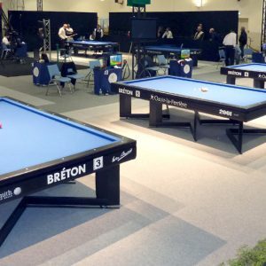 Una sala con varias mesas de billar, todas ellas etiquetadas como bretonas, ofrece un ambiente cl&aacute;sico de billar bret&oacute;n. Las mesas est&aacute;n ordenadas en un entorno de torneo bien iluminado, con algunas personas de pie observando al fondo.