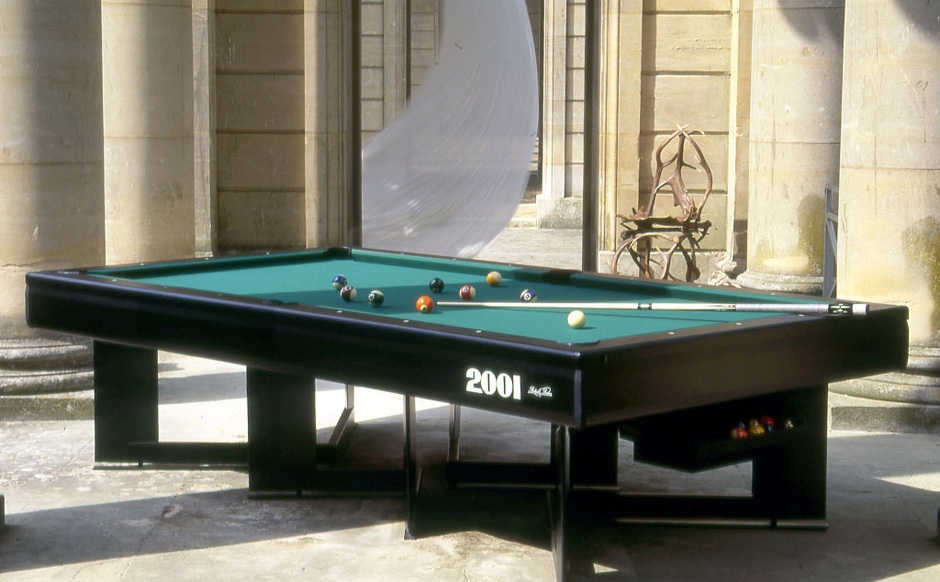 Une table de billard breton moderne avec une surface de jeu verte est installée dans une cour en pierre ensoleillée, entourée de hautes colonnes. Les boules de billard sont soigneusement disposées sur la table et une sculpture moderne et élégante se dresse gracieusement en arrière-plan.