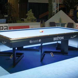 Una mesa de billar bien iluminada con dos bolas de billar en el centro se exhibe en un entorno de torneo. La mesa tiene la marca Bréton y está rodeada de carteles de patrocinadores. El lugar cuenta con una iluminación tenue con asientos adicionales en el fondo.