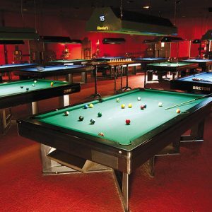Una sala de billar con poca luz y varias mesas de billar dispuestas en filas. Cada mesa está equipada con un juego de bolas de billar y las luces del techo iluminan las superficies de juego. La sala tiene un ambiente cálido y rojizo.