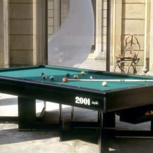 Una mesa de billar negra con fieltro verde adorna un patio de piedra iluminado por el sol, que recuerda a un juego de billar bretón. Varias bolas de billar se encuentran esparcidas por la superficie, mientras que grandes columnas de piedra y una escultura de metal única se yerguen orgullosas en el fondo.